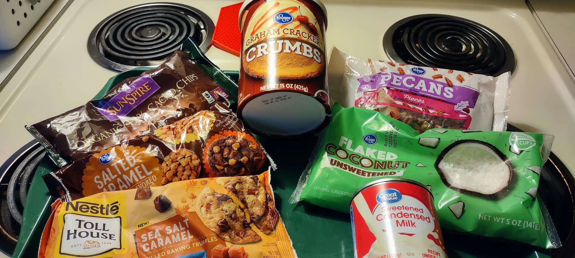 Seven-or-eight-layer cookie bar ingredients laying on a range: canister of graham cracker crumbs, bag of pecans, flaked/shaved coconut, can of sweetened condensed milk and bags of various flavors of chocolate baking chips.
