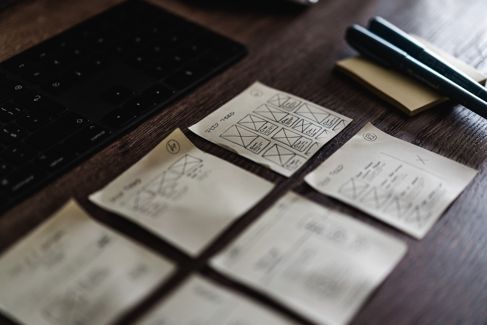 Close-up photo of various sticky notes showing various web layout designs next to some pens with a keyboard to the right, above them. They are on a dark brown wooden desk.